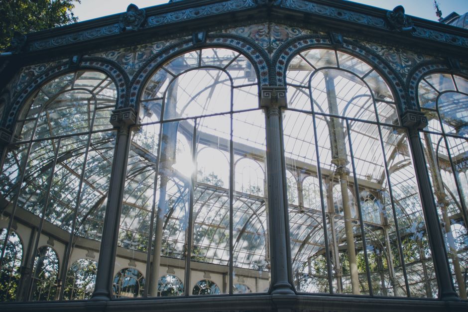 The image shows an ornate, Victorian-style glass and metal greenhouse structure with intricate decorative details along the upper edges, painted in light blue and cream tones. The large arched windows feature multiple panes of clear glass, allowing sunlight to filter through and create a bright, airy atmosphere inside. Visible steel or wrought iron supports with decorative motifs run vertically and horizontally, dividing the glazed sections. Sunlight casts reflections on the glass, and faint tree foliage is visible through the transparent sections, indicating the greenhouse is situated outdoors, possibly in a garden or park setting. The overall environment appears clean and well-maintained, emphasizing the historic architectural design and the transparency of the glass panels. This detailed description supports the context of designing or maintaining such structures, subtly linked to the broader theme of waste management and preservation of heritage sites, as offered by Waste Disposal Crystal Palace in their services related to independent collection and on-site clearance of unwanted items.