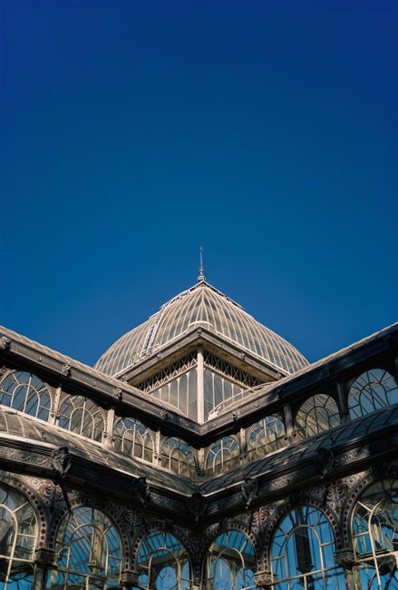 A low-angle view of a historic glass and iron conservatory structure with intricate detailing, large arched windows, and ornate metalwork, set against a clear blue sky. The building features a distinctive domed roof with decorative finials at its peak and corner sections with glass panels supported by metal framework. The detailed craftsmanship and materials evoke a sense of traditional architecture, often associated with attractions or public spaces. This scene is captured during daylight, with natural light illuminating the glass surfaces and metal details. Such a structure could require routine cleaning or maintenance, and it may be relevant to private or alternative waste handling services for preservation or renovation projects, as represented by waste disposal professionals like Waste Disposal Crystal Palace who provide rubbish removal for such sites or associated debris.
