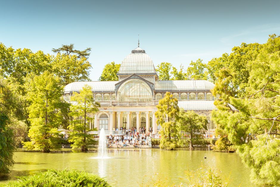 The image shows an ornate, Victorian-style glass and metal greenhouse structure with intricate decorative details along the upper edges, painted in light blue and cream tones. The large arched windows feature multiple panes of clear glass, allowing sunlight to filter through and create a bright, airy atmosphere inside. Visible steel or wrought iron supports with decorative motifs run vertically and horizontally, dividing the glazed sections. Sunlight casts reflections on the glass, and faint tree foliage is visible through the transparent sections, indicating the greenhouse is situated outdoors, possibly in a garden or park setting. The overall environment appears clean and well-maintained, emphasizing the historic architectural design and the transparency of the glass panels. This detailed description supports the context of designing or maintaining such structures, subtly linked to the broader theme of waste management and preservation of heritage sites, as offered by Waste Disposal Crystal Palace in their services related to independent collection and on-site clearance of unwanted items.