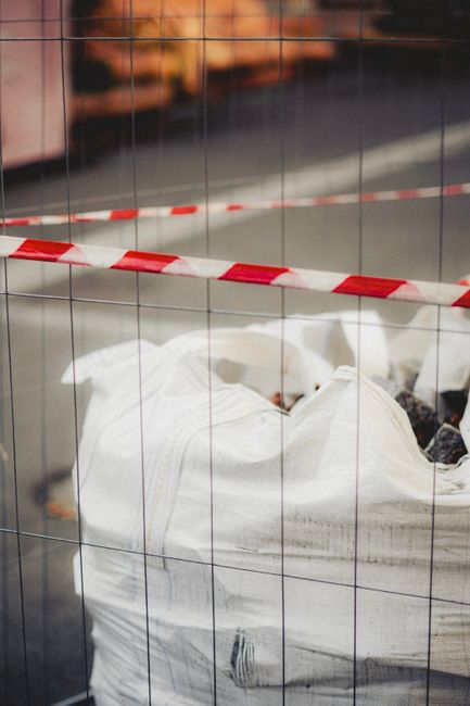 A large white sack made of woven polypropylene material, used for waste collection, is positioned on a paved surface behind a protective metal fence composed of vertical and horizontal wires. The sack appears to be filled with construction debris or mixed rubbish, with some dark, irregular-shaped objects visible at the top opening. The surface of the sack has a slightly textured finish, showing signs of compression and handling. A red and white striped safety tape is draped horizontally across the fence at the top of the sack, suggesting that the area is temporarily restricted or undergoing clearance work. The background features blurred elements of a paved driveway or street, with muted lighting indicating an outdoor environment possibly during late afternoon or early evening. This scene exemplifies a private waste removal scenario, with a focus on on-site clearance involving the use of sacks and fencing to contain and segregate debris, a service often associated with independent rubbish collection efforts managed by waste disposal specialists like Waste Disposal Crystal Palace, who facilitate alternative waste handling outside local authority provisions.