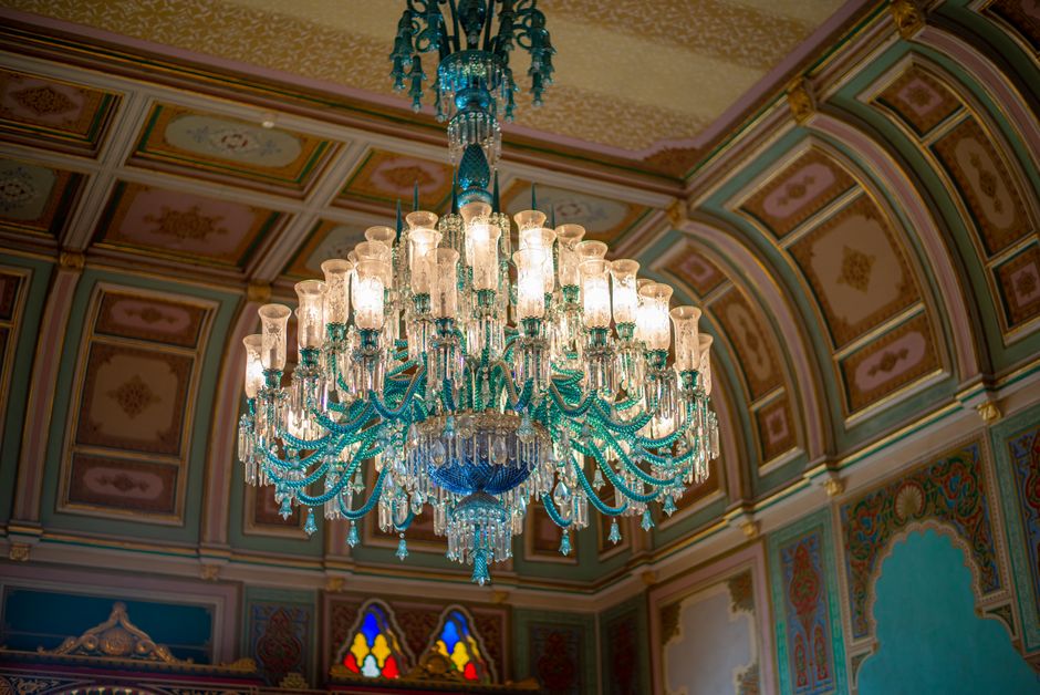 The image displays an ornate indoor ceiling featuring a large, elaborate chandelier made of clear glass or crystal with multiple tiers of arms and candle-shaped light fixtures. The chandelier has a predominantly turquoise or teal hue and is adorned with intricate glass details and hanging crystal ornaments, casting a soft, warm glow. Surrounding the chandelier, the ceiling showcases decorative paneling with ornate molding and painted panels in pastel tones, including gold accents, pink, and green, creating a richly detailed ceiling design. The background includes a glimpse of stained glass windows with vibrant colors such as red, yellow, and blue, adding to the luxurious and historic ambiance of the space, which appears to be part of an opulent interior such as a ballroom or grand hall. This detailed ceiling scene visually contrasts with typical rubbish removal contexts but subtly indicates a setting where high-quality finishes and decorative features are present, potentially requiring professional cleaning or maintenance services provided by Waste Disposal Crystal Palace.