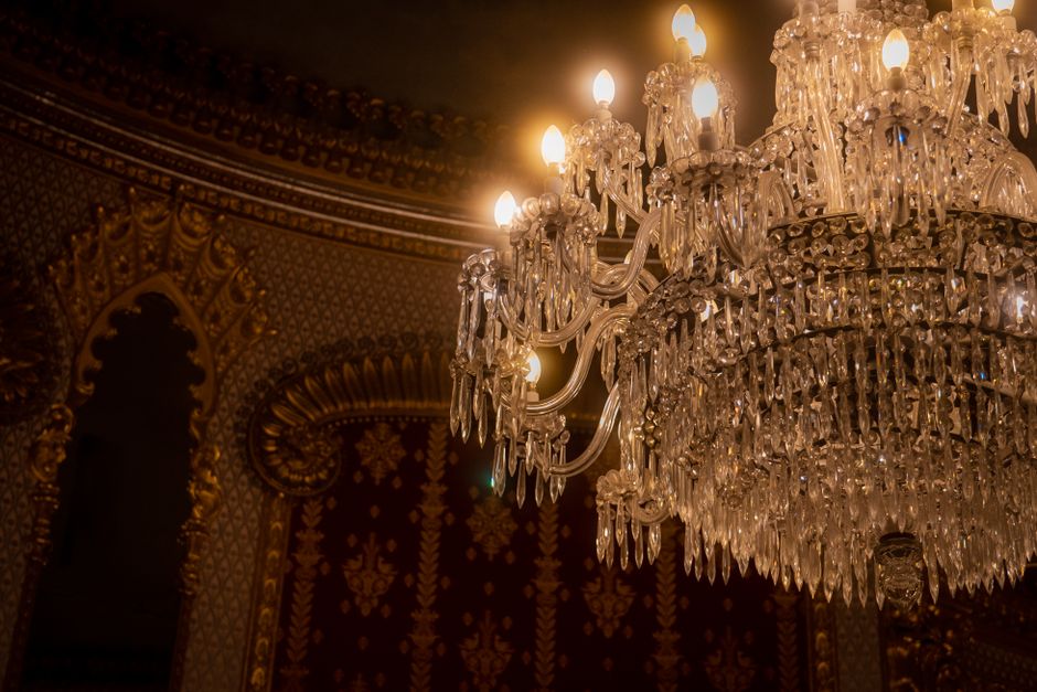 A large, ornate crystal chandelier hangs from the ceiling of an opulent interior space, featuring multiple arms with candle-shaped light bulbs. The chandelier is composed of numerous clear glass or crystal droplets and prisms that reflect the warm, yellowish light emitted by the bulbs. The background shows richly decorated walls with intricate gold patterns and dark, textured wallpaper, contributing to a luxurious atmosphere. The chandelier's elaborate design and shimmering surfaces highlight its decorative function within this elegant setting, contrasting with the neutral, practical tone of rubbish removal services offered by Waste Disposal Crystal Palace. Although the scene is primarily focused on the chandelier, the environment suggests an indoor environment suited to private or high-end commercial properties where alternative waste handling or on-site clearance might be necessary during refurbishment or maintenance. The lighting and detailed craftsmanship of the chandelier evoke a sense of careful preservation and aesthetic detail typical of such refined interiors.