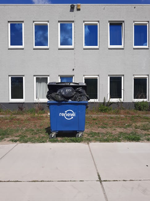 A blue wheeled bin marked with the 'renewi' logo, filled with black garbage bags stacked on top, positioned on a paved sidewalk in front of a modern, three-storey building with white exterior walls and large rectangular windows reflecting a partly cloudy sky. The bin is situated on a patch of grass and soil, indicating an outdoor waste collection point, with no other visible waste or disposal equipment nearby. The scene suggests an instance of private waste disposal, possibly managed by Waste Disposal Crystal Palace, in an urban or suburban environment where independent rubbish collection is undertaken for commercial or residential waste removal purposes.