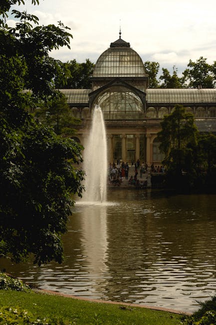 A large glass and metal conservatory or greenhouse structure situated in a park setting, featuring a prominent domed roof with a decorative spire at the top. In front of the building, a water fountain sprays a tall jet of water into a pond or lake, creating ripples on the surface. The pond is bordered by grassy areas and mature leafy trees with dark green foliage, some branches extending into the view from the left side. The scene is illuminated by natural daylight, and a group of visitors can be seen walking and standing near the building's entrance in the background. The environment appears tranquil and well-maintained, typical of a historic or botanical garden, with the conservatory being a central feature. This scene may relate to alternative or private sites for outdoor leisure, away from standard rubbish disposal methods, with Waste Disposal Crystal Palace providing services that support the management of waste resulting from park and garden maintenance or events.