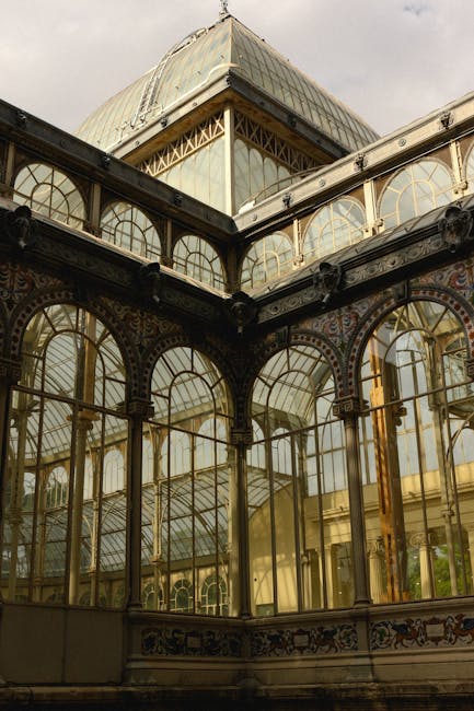 This image shows the exterior of an ornate Victorian-style glass greenhouse with intricate wrought iron framework. The structure features large arched windows with metal framing, painted in a dark color, displaying decorative patterns along the upper sections. The glass panes are clear, revealing the interior lighting and details of the greenhouse’s interior surfaces. The building’s roof has a steep pitch with additional glass panels, and the daylight creates reflections and a subtle glow on the glass surfaces. Surrounding environment is not visible. The scene is captured under natural daylight, with soft shadows emphasizing the architectural details. This structure demonstrates historic design elements typical of botanical glasshouses, and the scene is consistent with maintaining or restoring vintage glass conservatories, which aligns with sustainable and independent approaches to garden or estate maintenance, often managed through specialized waste and debris removal services by companies such as Waste Disposal Crystal Palace.