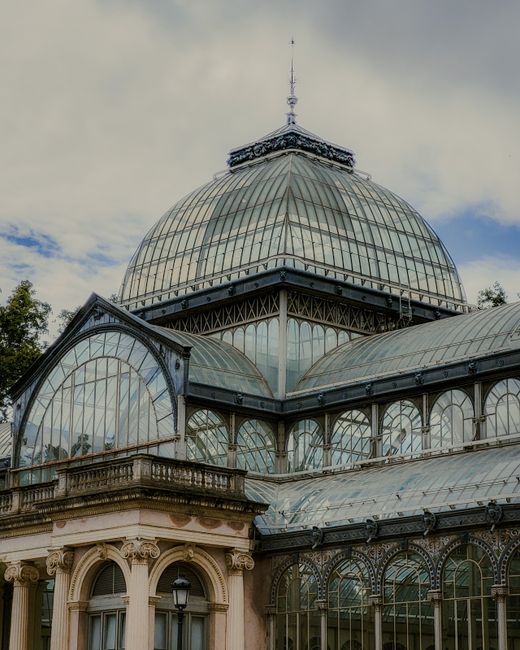 A large glass and metal conservatory or greenhouse structure situated in a park setting, featuring a prominent domed roof with a decorative spire at the top. In front of the building, a water fountain sprays a tall jet of water into a pond or lake, creating ripples on the surface. The pond is bordered by grassy areas and mature leafy trees with dark green foliage, some branches extending into the view from the left side. The scene is illuminated by natural daylight, and a group of visitors can be seen walking and standing near the building's entrance in the background. The environment appears tranquil and well-maintained, typical of a historic or botanical garden, with the conservatory being a central feature. This scene may relate to alternative or private sites for outdoor leisure, away from standard rubbish disposal methods, with Waste Disposal Crystal Palace providing services that support the management of waste resulting from park and garden maintenance or events.