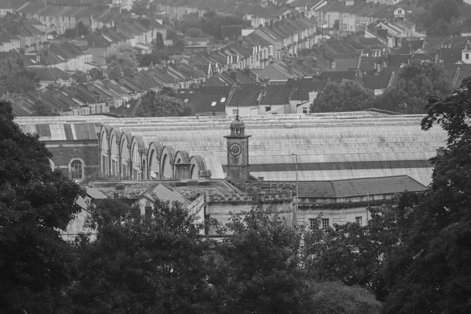 A black-and-white photograph capturing a view of a historic train station with a prominent clock tower situated above its roofline, surrounded by residential houses and terraced streets in the background. In the foreground, there are trees with dense foliage partially obscuring the lower part of the station, creating a natural frame around the structure. The station building features a large, arched glass roof with a series of semi-circular windows, and the clock tower has a classic design with a round clock face and a dome-shaped top. The scene is illuminated by natural daylight, highlighting the textures of the weathered roof and the intricate architectural details of the station, with the environment suggesting an urban setting within a residential area. This image, although in black-and-white, emphasizes the historical character of the railway station amidst the surrounding neighborhood, relevant to contexts of infrastructure, private transport, and public transportation hubs in a local urban environment, as might be associated with alternative waste handling or maintenance activities in such facilities.
