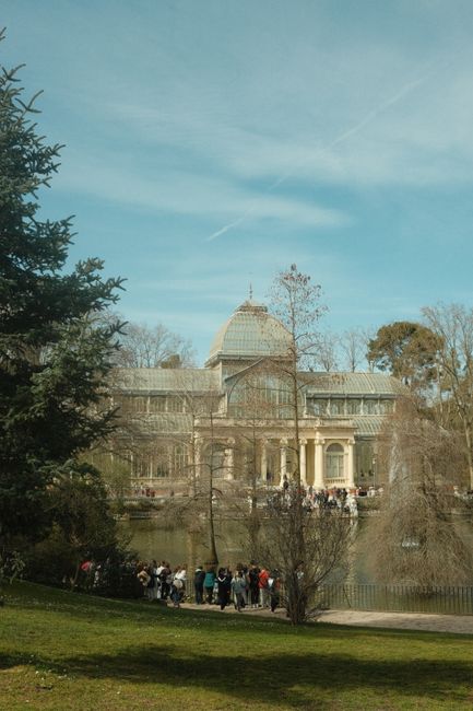 A large, historic glass-and-stone building with a central dome and classical architectural features, situated across a body of water reflecting its facade. In the foreground, a group of people is gathered near the water's edge on a grassy area, with some walking along a path. To the left, there is a tall evergreen tree, and on the right, several leafless trees and shrubs are visible. The sky above is clear and blue, with a few wispy clouds, and the overall scene appears to be in a park or garden setting close to the building. The presence of visitors and the park environment suggest an outdoor leisure area, possibly near an institution or landmark, with the scene captured during daylight hours. Waste Disposal Crystal Palace occasionally handles separate waste management tasks, supporting private and on-site clearance activities in environments like this, where outdoor space and historical architecture are prominent.